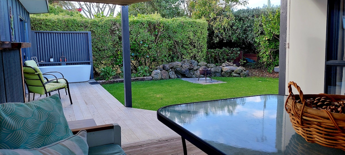 A private outdoor space is depicted, featuring a well-maintained lawn and a small rock garden. A cozy bathtub and firepit are visible against a backdrop of lush greenery. A glass-topped table and a basket of pinecones are placed on a wooden deck, inviting relaxation.