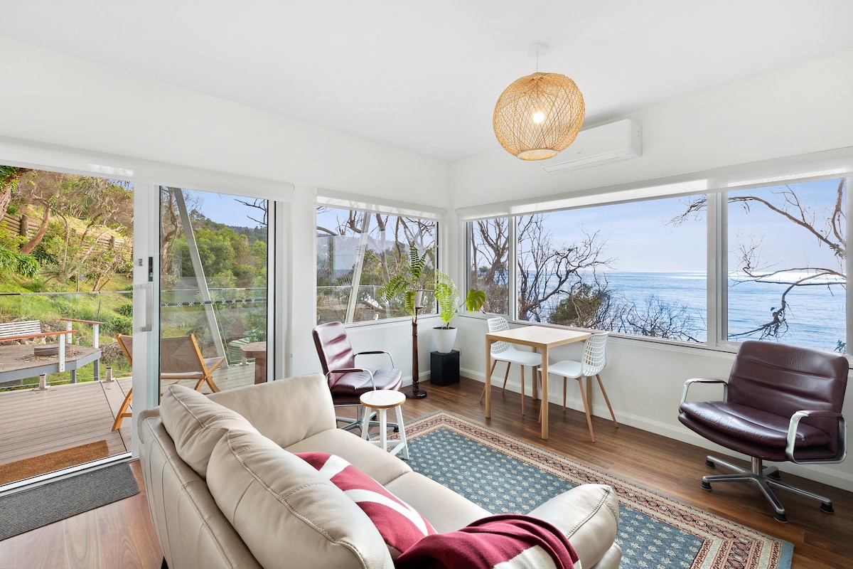 A cozy living area features large windows offering expansive views of the ocean and surrounding greenery. A comfortable sofa, two chairs, and a dining table create a welcoming space. Natural light fills the room, complemented by a pendant light overhead, enhancing the serene atmosphere.