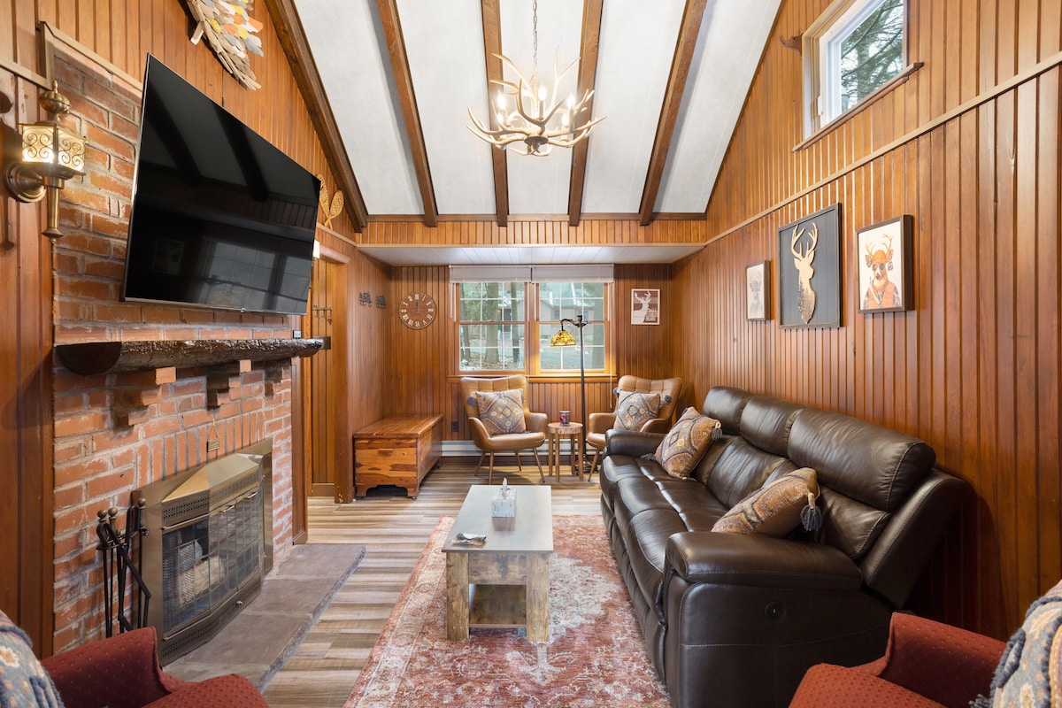 A warm living area features wood-paneled walls accented by exposed beams. A large flat-screen TV is mounted above a brick fireplace. Comfortable seating is arranged around a coffee table, with natural light streaming in through windows adorned with art. Decorative pieces add a rustic charm.