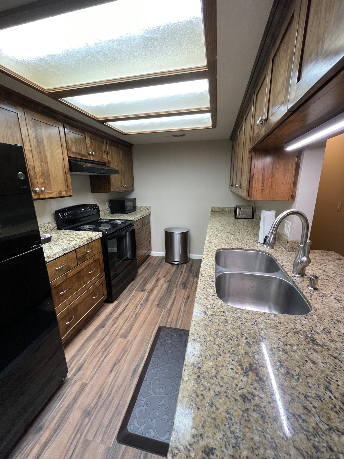 A fully equipped kitchen is presented with rich wood cabinetry and stainless steel appliances. The countertops are made of polished granite, providing ample space for meal preparation. Natural light filters through a skylight above, and a small waste bin is positioned near the double sink.