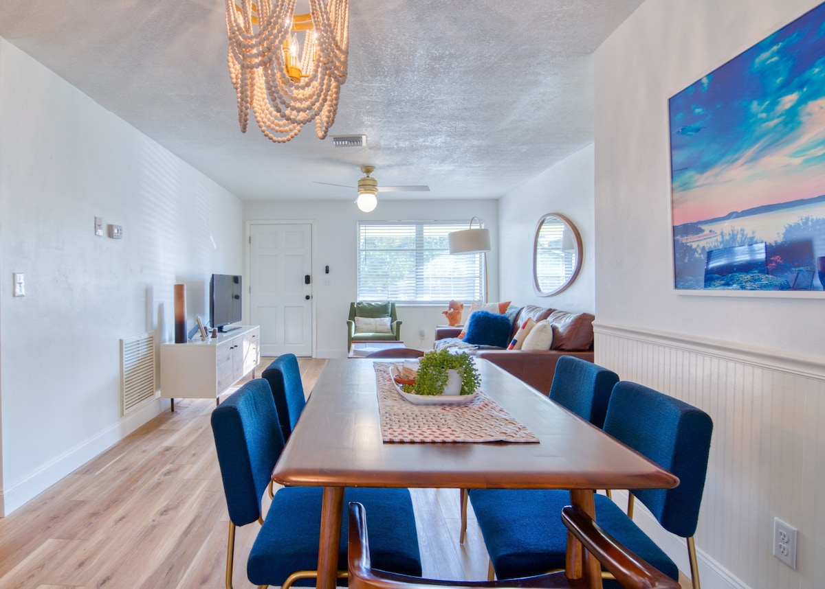 A spacious living and dining area is presented, featuring a wooden dining table surrounded by six blue chairs. Natural light enters through large windows, highlighting the soft tones of the decor. A comfortable seating area with a sofa and television is visible in the background.