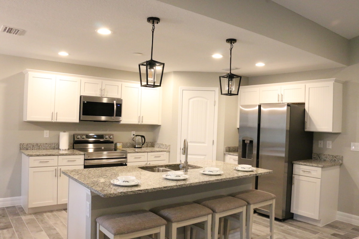 A modern kitchen features a spacious island with seating for four. Stainless steel appliances complement the light cabinetry, and granite countertops provide ample workspace. Pendant lights hang above the island, illuminating the area. The room is designed with an open layout, enhancing functionality.