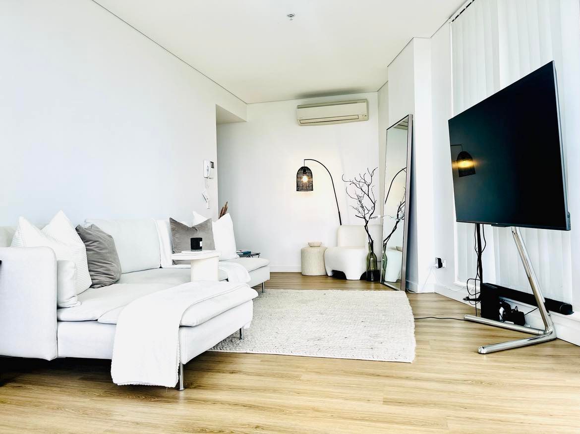 A bright living area features a spacious sofa paired with decorative cushions, complemented by a soft rug and a modern flat-screen TV. Natural light floods the space, accentuated by minimalist decor that includes a floor lamp and a decorative plant.