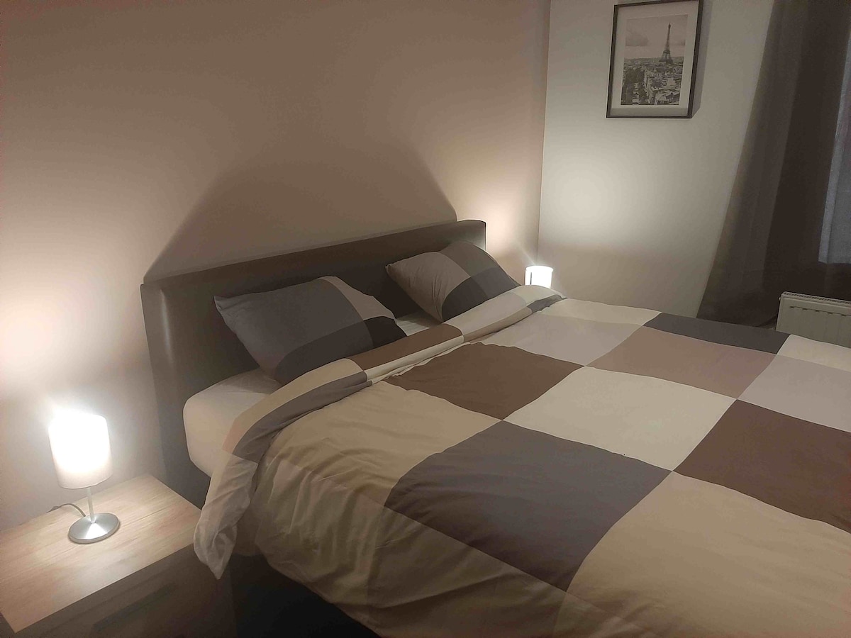 A spacious bed is dressed in a patchwork comforter with shades of beige, brown, and gray. Soft lighting from bedside lamps creates a gentle glow, complementing the neutral color scheme. A framed picture is displayed on the wall, adding a touch of decor.