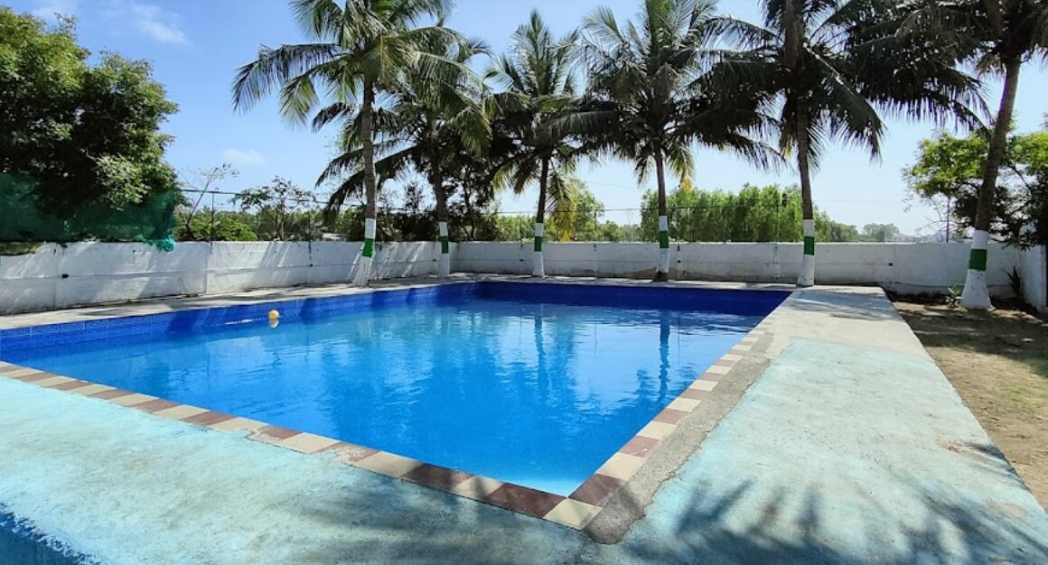A clear swimming pool is surrounded by palm trees and lush greenery. The pool features a tiled border and is framed by a concrete deck, providing ample space for relaxation. Bright blue sky enhances the vibrant colors of the scene.