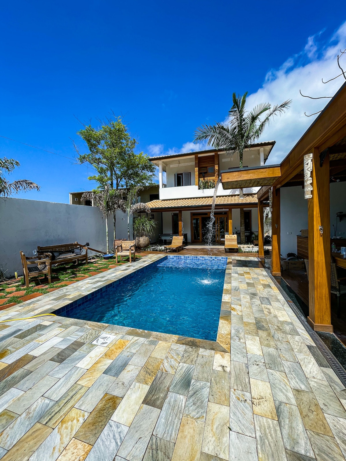 A refreshing pool is surrounded by natural stone tiles and lush greenery, with comfortable seating positioned nearby. The two-story house, featuring a blend of modern and traditional architecture, is visible in the background under a clear blue sky.