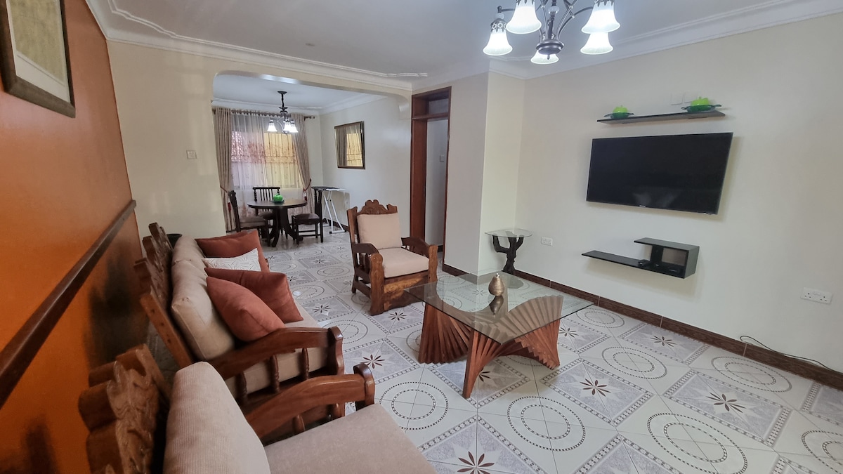 A spacious living area is illuminated by overhead lighting, featuring wooden furniture and comfortable seating. A glass coffee table is centered on a patterned floor. A mounted TV is visible on the wall, while a dining table can be seen in the background.