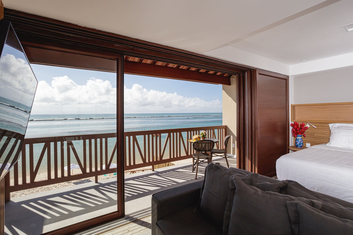 A master suite features a spacious king bed and a balcony with an unobstructed view of the calm sea. Large glass doors open to the outdoor space, while a comfortable sofa and a smart television complete the inviting atmosphere.