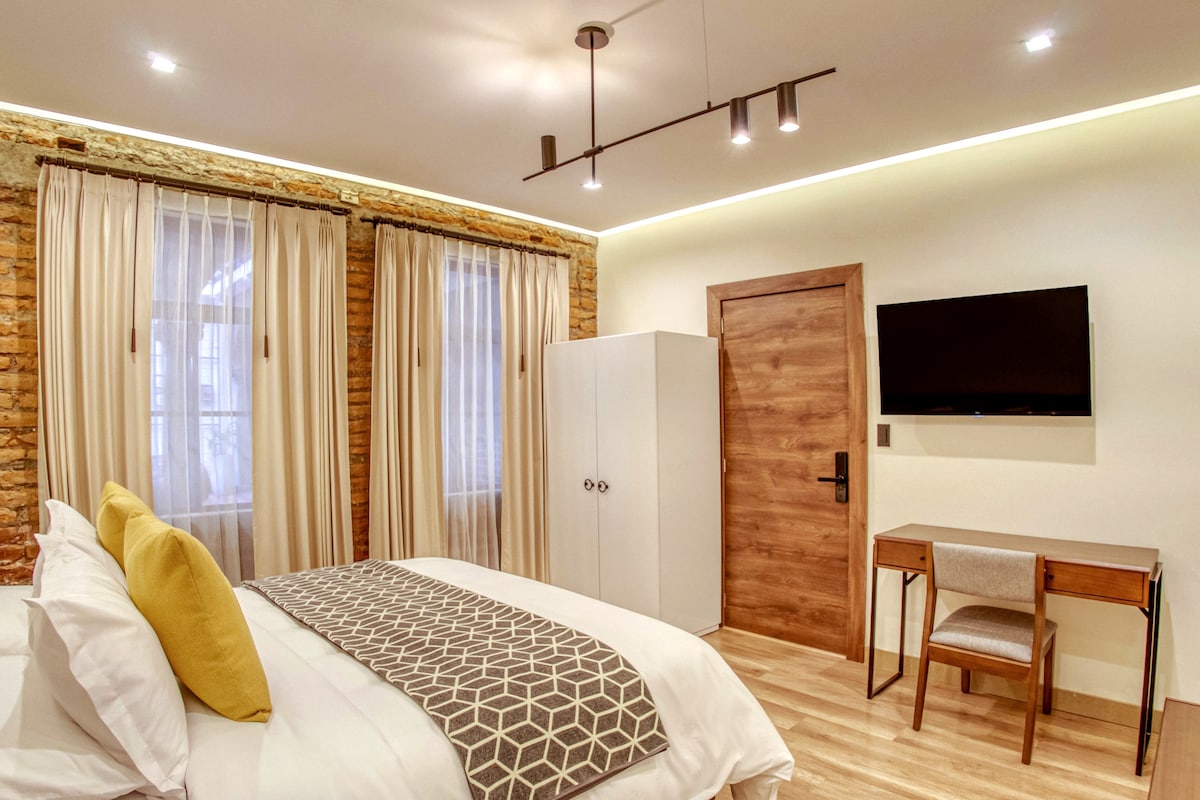 A well-appointed room features an inviting bed with decorative pillows. Exposed brick walls contrast with modern furnishings, including a sleek desk and a flat-screen television. Natural light filters through sheer curtains, enhancing the room's bright and airy ambiance.