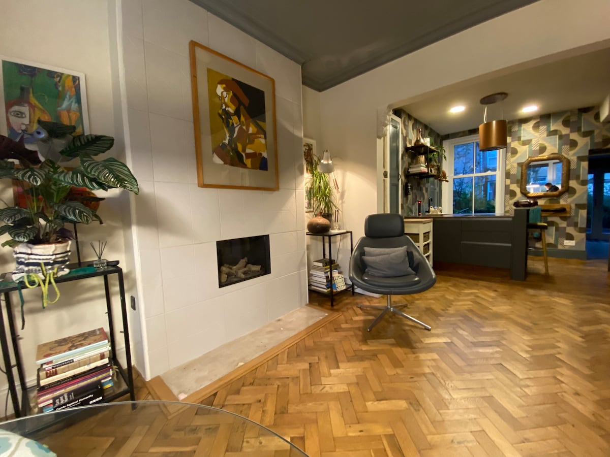 A stylish living area features a sleek chair positioned near a minimalist fireplace. A colorful artwork adorns the white wall, complemented by a leafy plant on a nearby stand. Warm wooden flooring enhances the inviting environment, leading to an adjacent kitchen space.