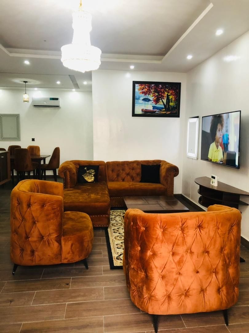 A modern living area features plush, orange tufted furniture, including a spacious sectional couch and two accent chairs. A large television is mounted on the wall, and a colorful artwork is displayed above a decorative table. Soft lighting from the ceiling adds a welcoming touch.