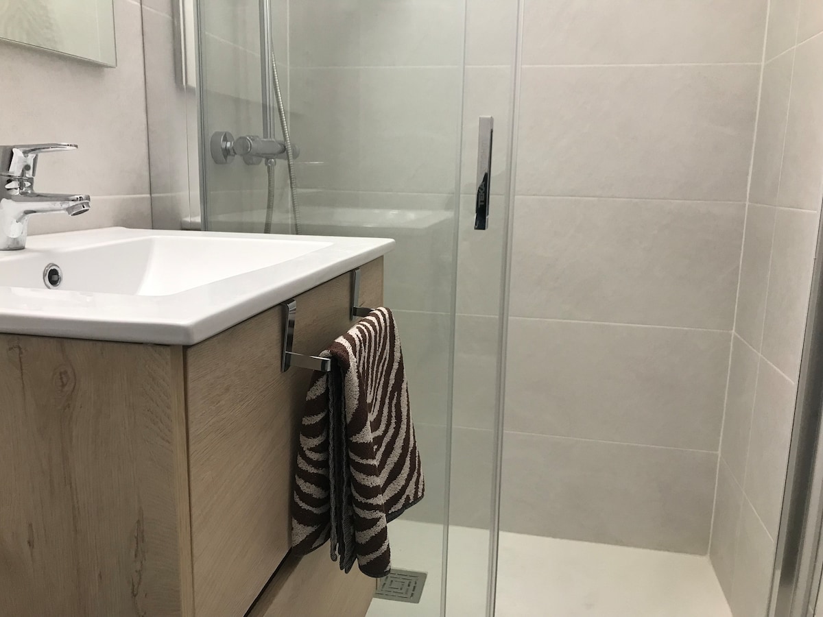 A modern bathroom is presented with a clear glass shower enclosure and sleek white tiles. Light wood cabinetry complements the space, with a white countertop featuring a sink. A striped towel hangs elegantly from the cabinet, adding a touch of texture.