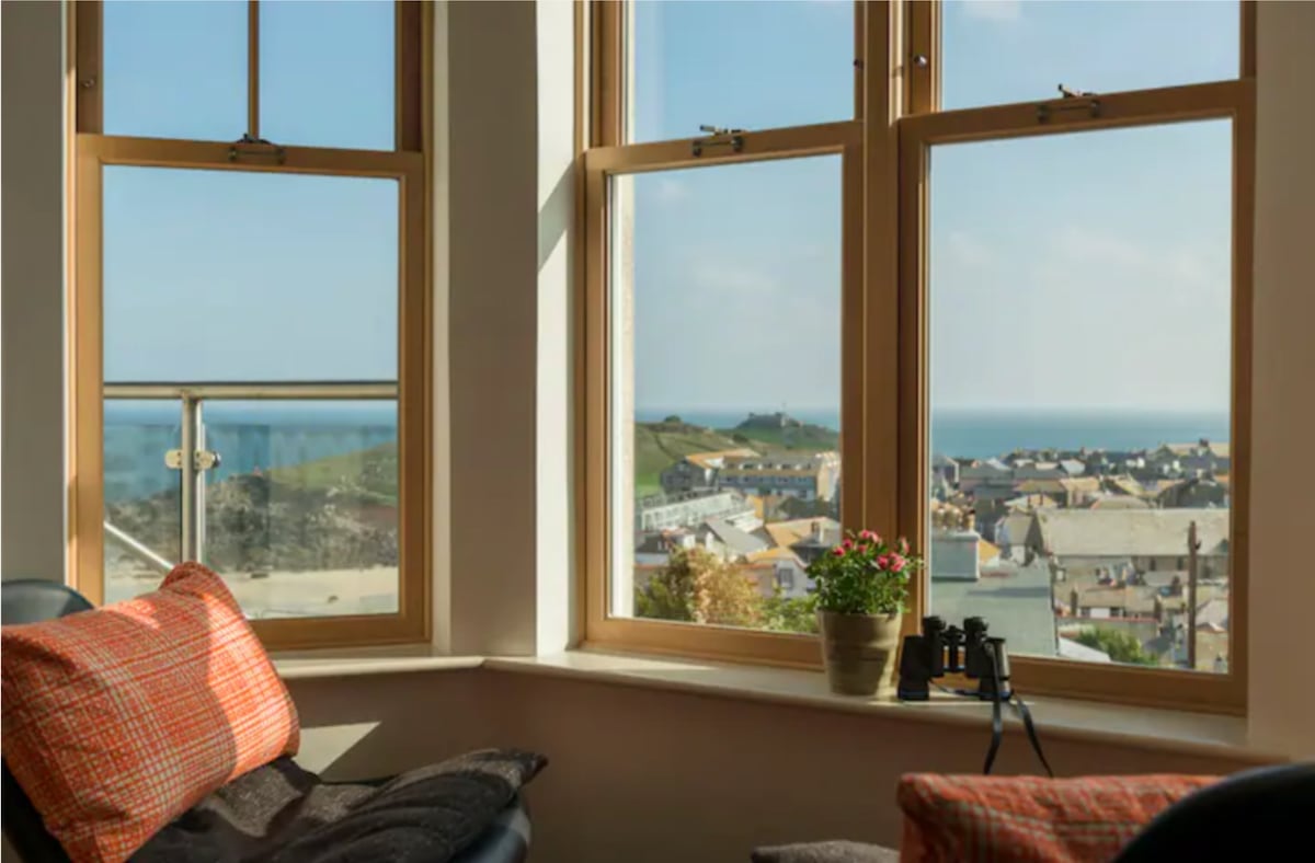 Large bay windows frame coastal views of St Ives, showcasing the sea and town. A small potted plant sits on the windowsill beside a pair of binoculars, enhancing the space. Comfortable seating with textured cushions invites relaxation while enjoying the scenery.
