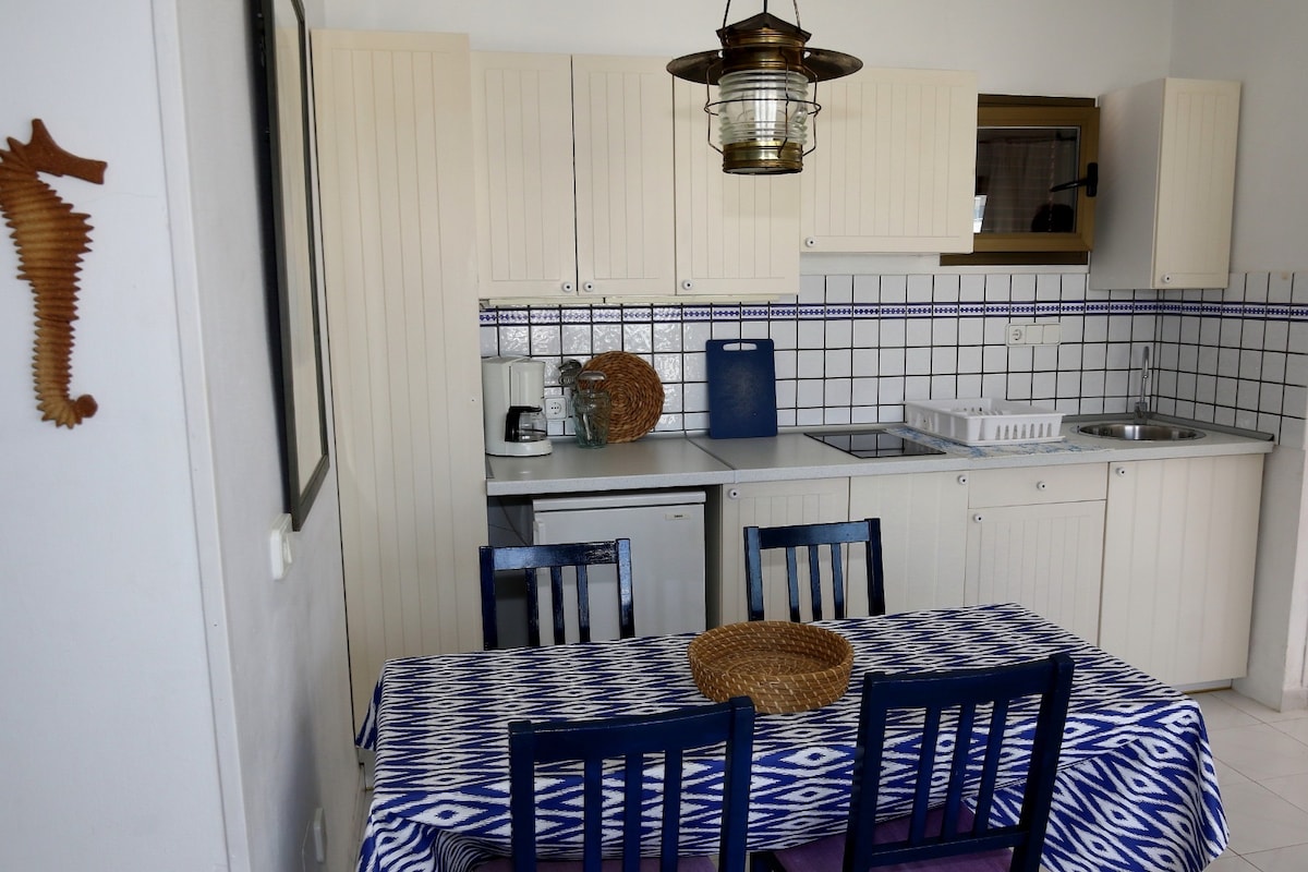 A functional kitchen space is presented, featuring white cabinetry and a tiled backsplash. A dining table with blue chairs is set beneath a pendant light. Appliances, including a refrigerator and stove, are integrated into the design, offering a practical cooking area.