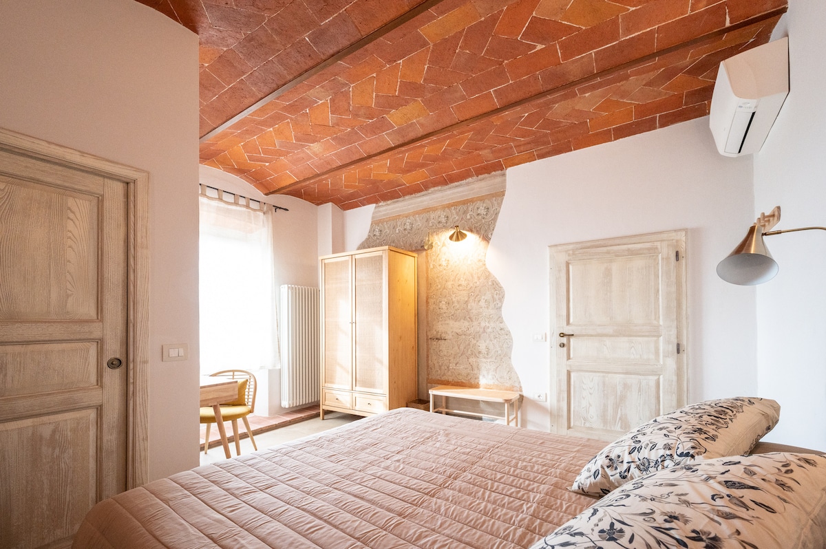 A spacious bedroom features a warm terracotta ceiling with herringbone patterns. Natural light streams in through a window, illuminating the wooden furniture, including a wardrobe and a small table. Air conditioning is also visible, ensuring a comfortable environment.