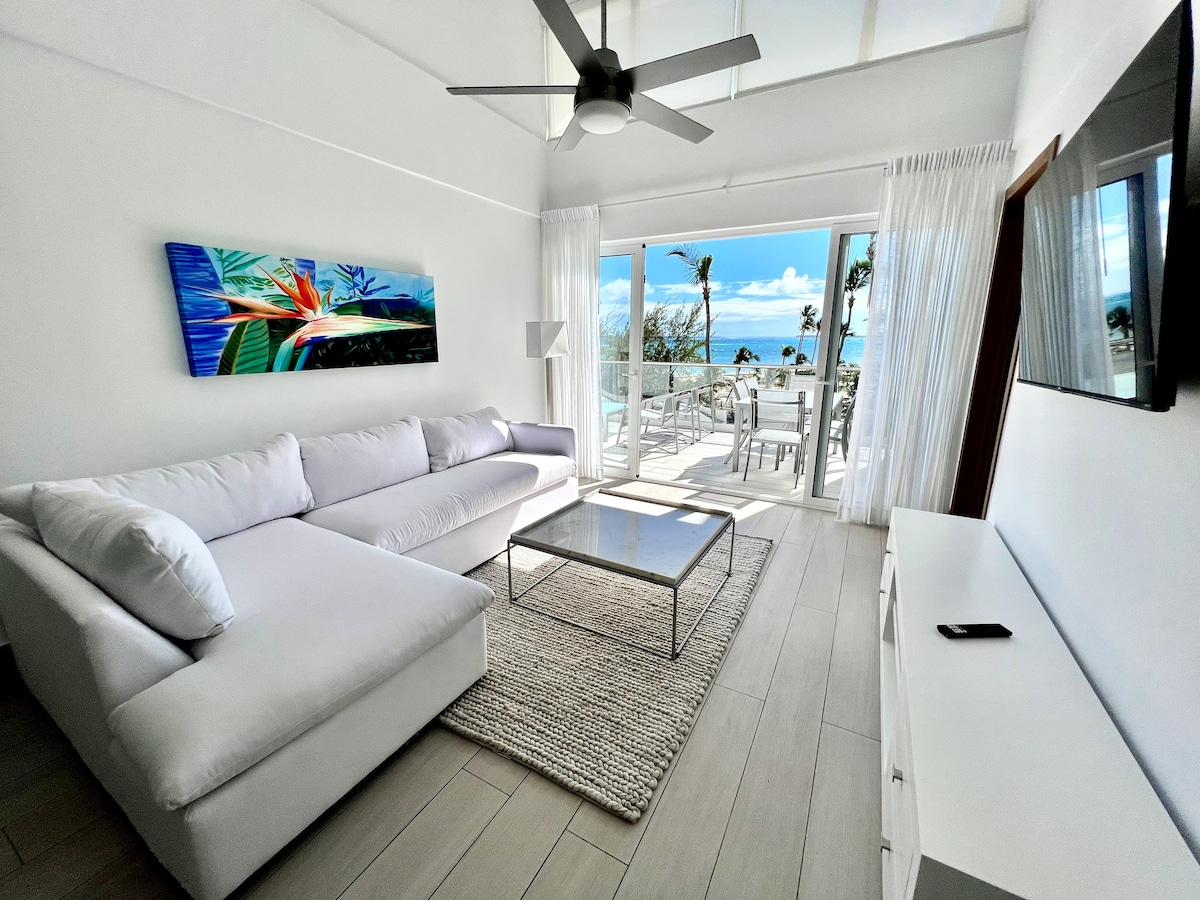 A bright living area features a spacious white sectional sofa paired with a glass coffee table. Light-colored walls and flooring create an airy feel. Large sliding glass doors lead to a terrace with sea views, while a colorful artwork adds a vibrant touch to the room.