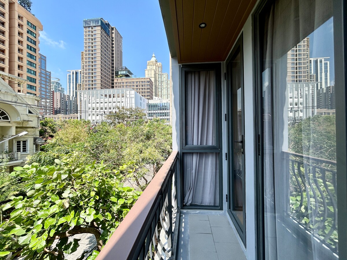 A private balcony is presented with a view of surrounding greenery and city skyscrapers, allowing for fresh air and natural light. The balcony features a railing and is accessed through glass doors that lead into the living space.