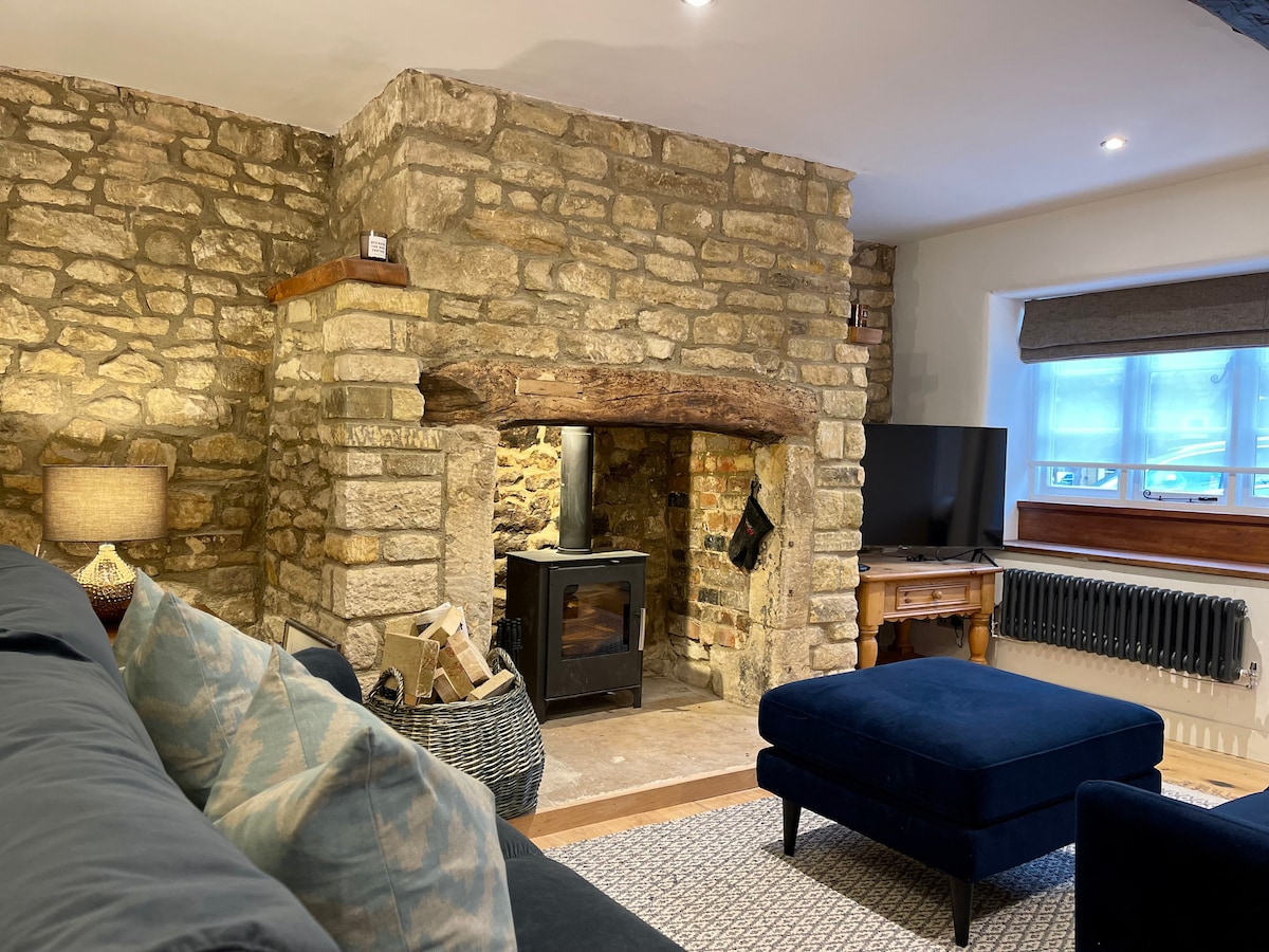 A cozy living area features a Cotswold stone wall with a rustic log burner, providing a warm focal point. Soft furnishings include a dark blue ottoman and a sectional sofa adorned with textured cushions. Natural light filters through a nearby window, illuminating the space.