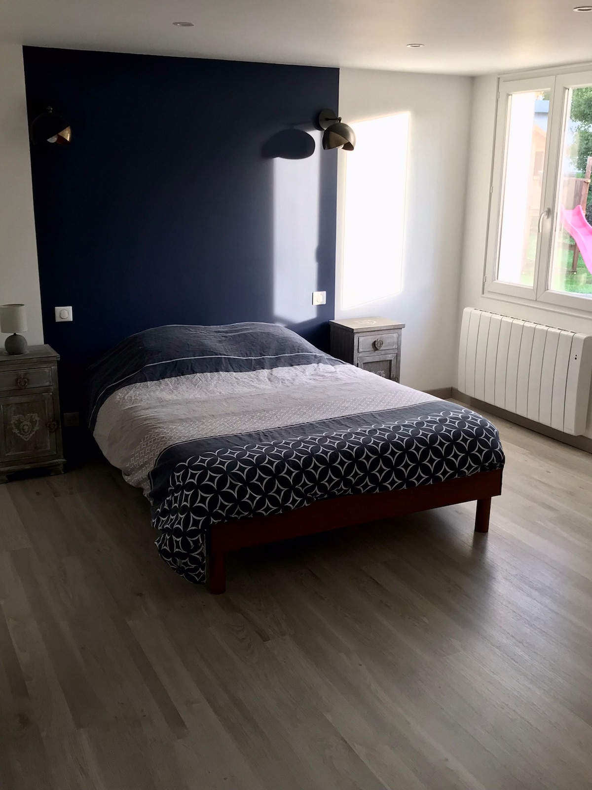 A cozy bedroom features a spacious bed with a patterned blanket. Wall sconces provide soft lighting, while a large window allows natural light to fill the room. Nightstands are placed on either side of the bed, contributing to the room's simplicity and functionality.