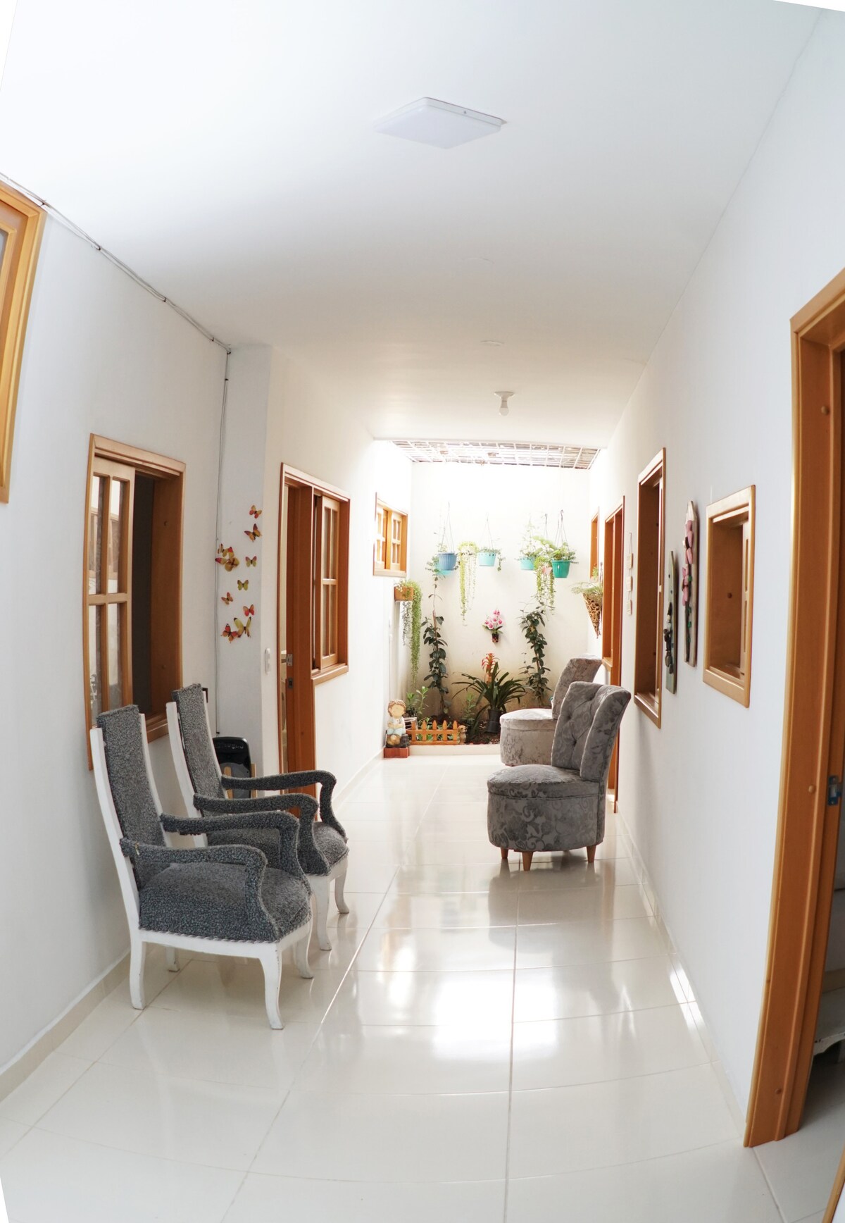A well-lit hallway features a glossy tiled floor and white walls adorned with framed mirrors. Two windows allow natural light to enter, while plants are displayed at the end. Comfortable chairs are arranged along one side, creating an inviting space for relaxation.