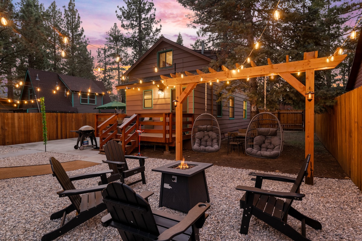 An outdoor space features a gravel area with four black Adirondack chairs arranged around a fire pit. A wooden pergola with string lights provides ambiance. In the background, the cozy cabin is visible, flanked by trees and lit by the soft glow of twilight.