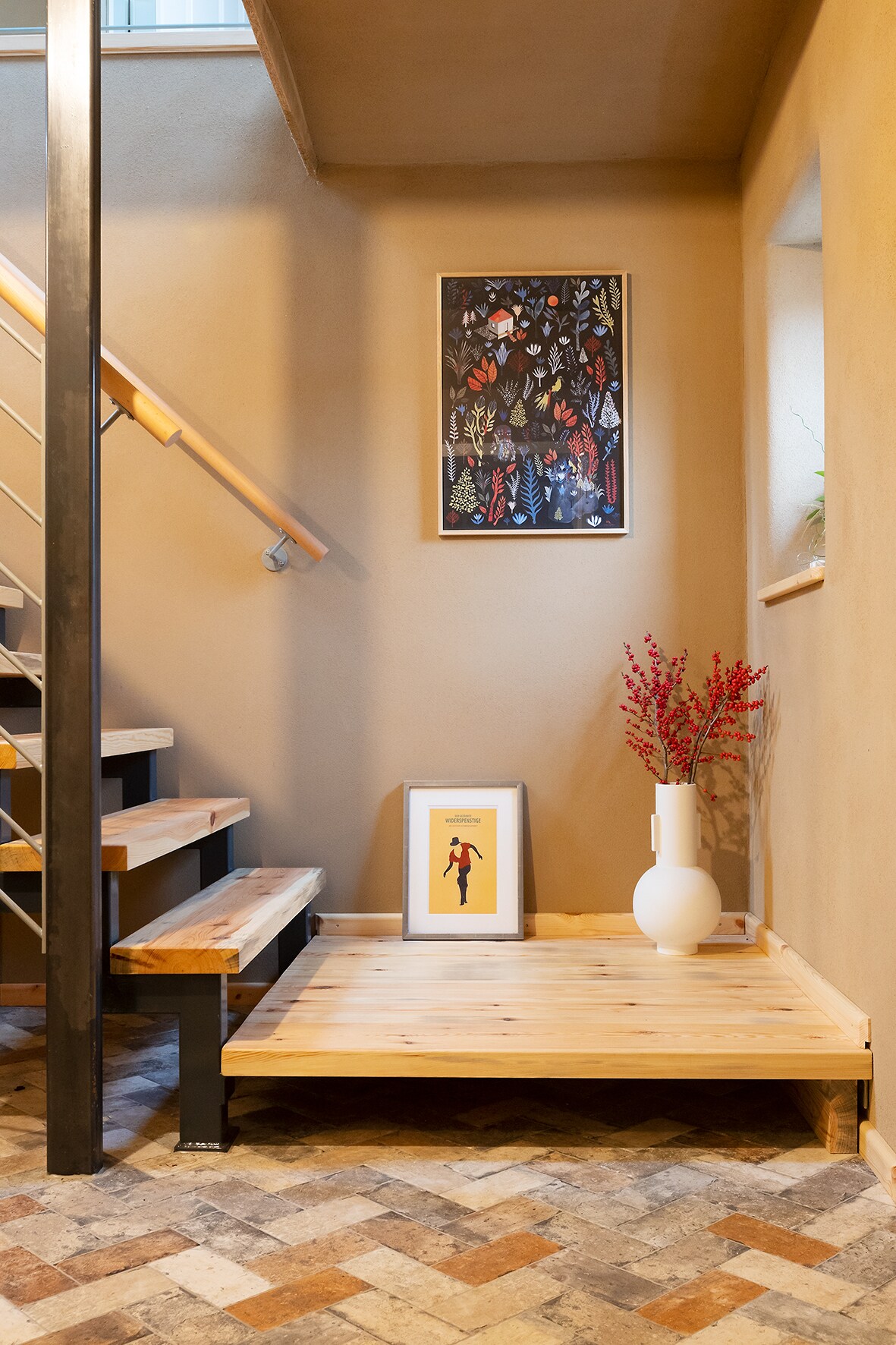 A welcoming entryway features a staircase with wooden steps and a metal railing. A piece of framed artwork adorns the wall, complemented by a vase with red flowers. A wooden platform provides space for decorative items, and a patterned floor adds texture to the area.