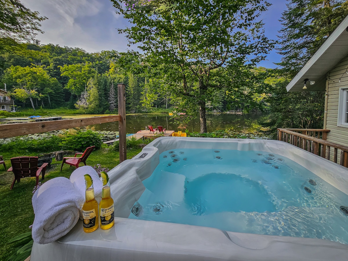 A hot tub is positioned on a wooden deck, with bubbles visible in the water. Soft towels are neatly arranged beside the tub, and drinks are placed on the side. Lush greenery and a serene lake view complete the tranquil setting.