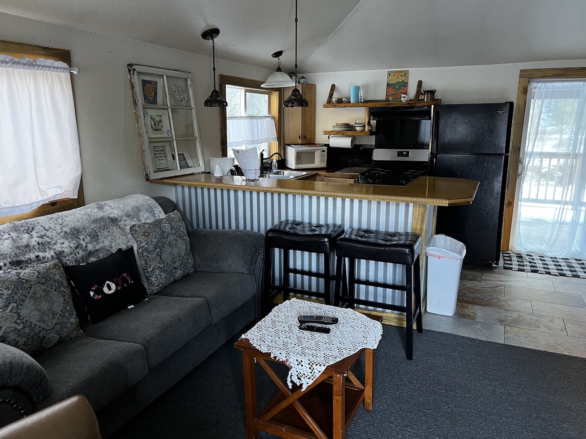 A living area is visible, featuring a comfortable couch and a wooden coffee table. Light streams in through the windows, illuminating the adjoining kitchen equipped with a microwave, stove, and refrigerator. Two stools are positioned at the countertop, enhancing a casual dining space.