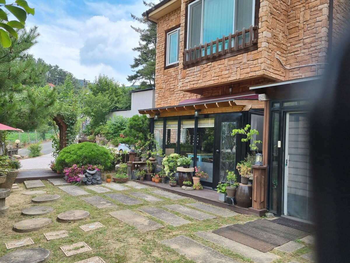 A stone-clad exterior of a two-story house is surrounded by vibrant greenery and flower pots. A charming pathway leads to the entrance, flanked by decorative plants and stone stepping stones. Large windows allow plenty of natural light to enter, creating an inviting atmosphere.