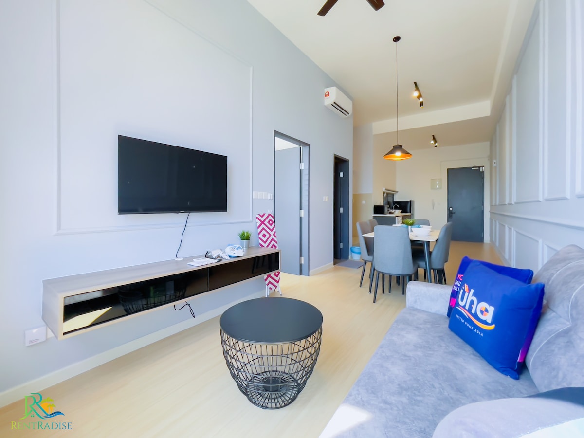 A modern living area features a sleek flat-screen TV mounted on the wall above a contemporary media console. A round black wire coffee table sits centrally, surrounded by a grey sofa and a dining table with four chairs. Bright light floods the space through large windows.