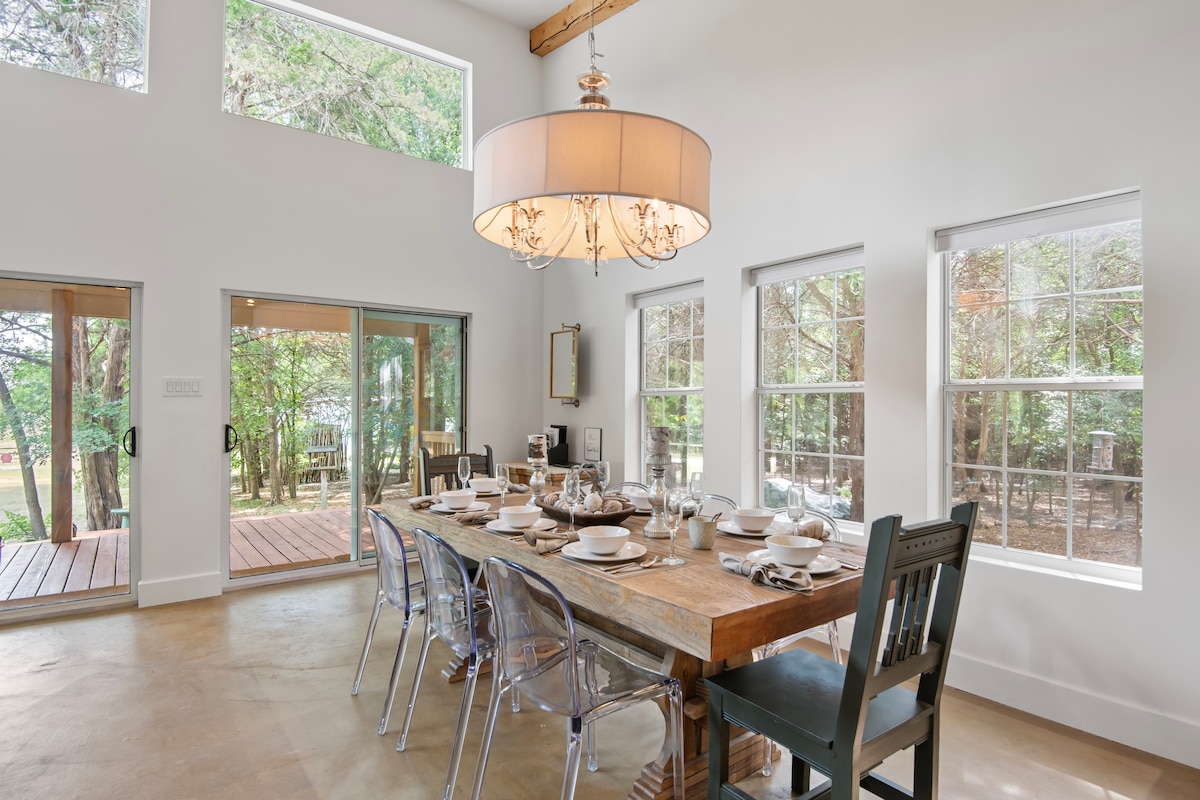 A well-lit dining area features a large wooden table set for a meal, surrounded by stylish ghost chairs. Windows on two walls provide views of the wooded surroundings, while a modern chandelier adds elegance to the space.