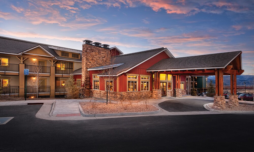 A welcoming resort entrance is showcased, featuring wooden beams and a stone facade. Large windows allow natural light to fill the lobby area. The surrounding landscape includes neatly arranged shrubs with a gravel path leading to the entrance under a colorful sky.