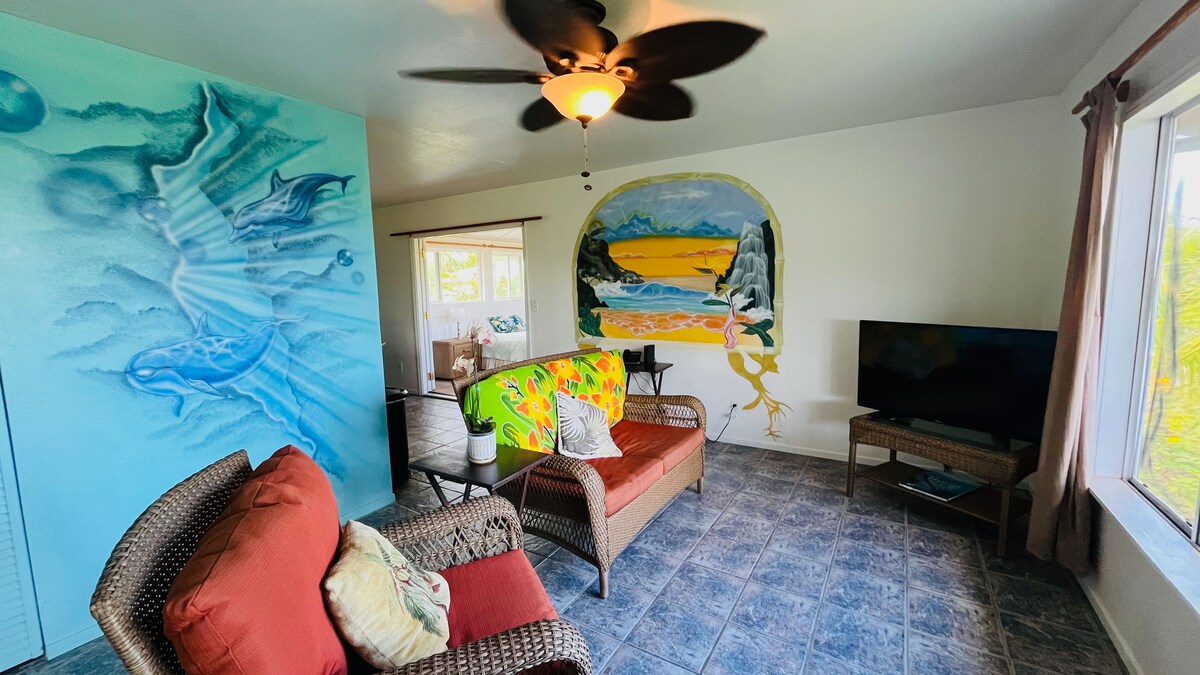 A cozy living area features a comfortable seating arrangement, with a sofa and chairs adorned with throw pillows. The walls display vibrant murals, one depicting a scenic landscape. Natural light enters through large windows, and a ceiling fan adds to the room's ventilation.