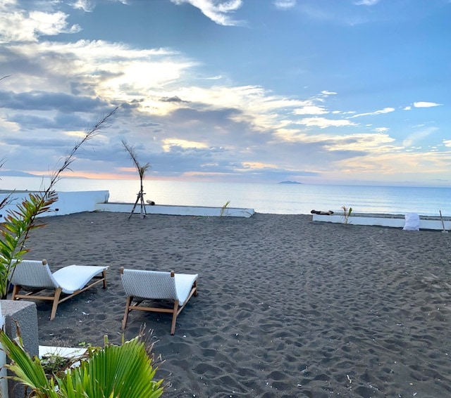 Two lounge chairs are positioned on a sandy area, facing a serene ocean view. The horizon features gentle waves and a soft sky transitioning from blue to pastel hues, while a few distant islands are visible on the horizon.