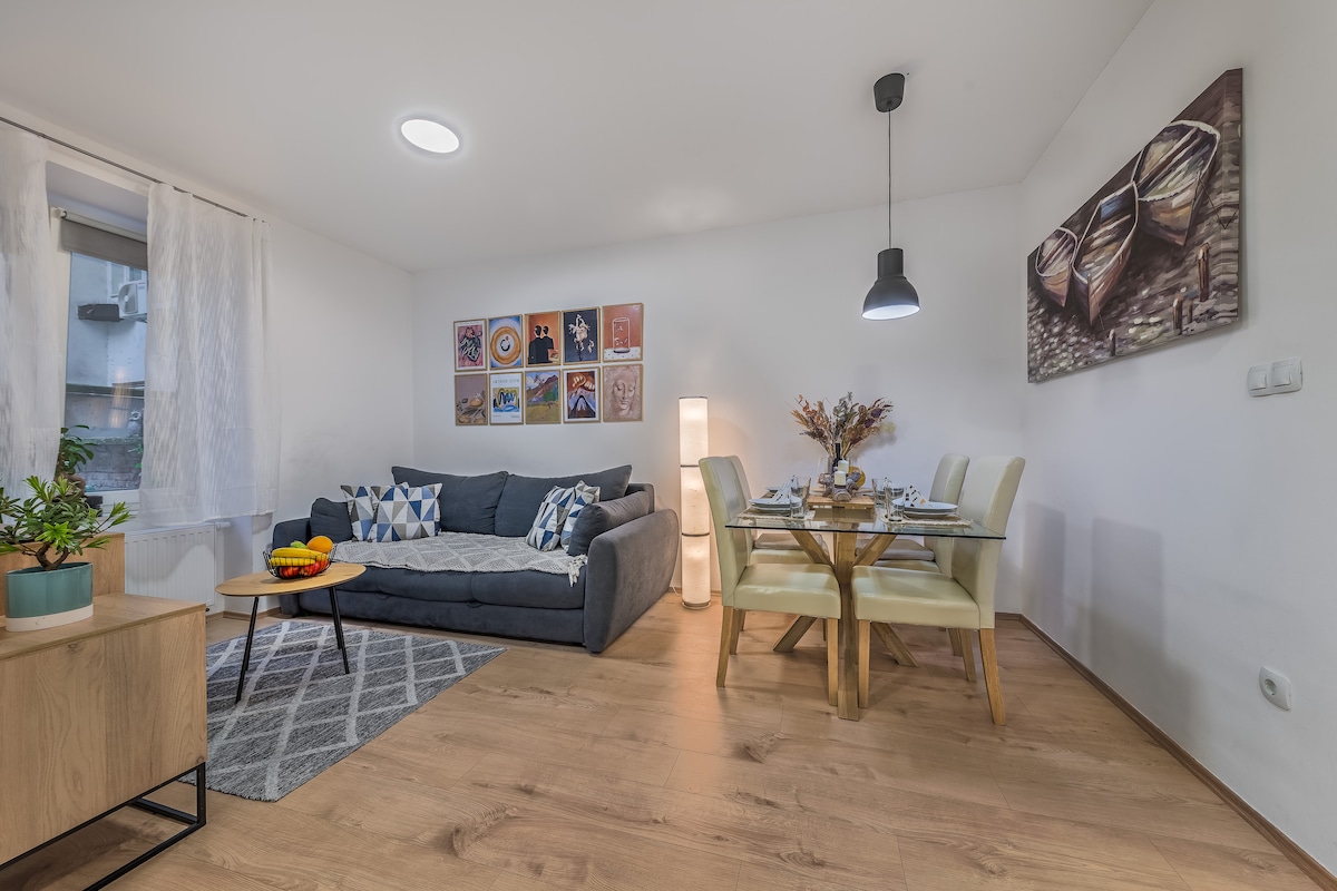 The living area features a spacious sofa adorned with decorative pillows, paired with a dining table set for six. Colorful artwork is displayed on the wall, and natural light enters through the window, complementing the warm tones of the wooden floor.