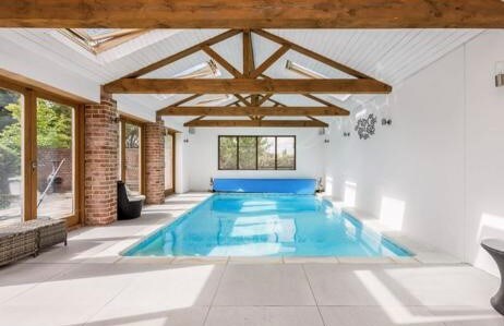 A bright indoor swimming pool is framed by large glass windows and a beamed ceiling, allowing natural light to illuminate the space. A serene blue water surface reflects the surrounding walls, while comfortable seating is placed nearby for relaxation.