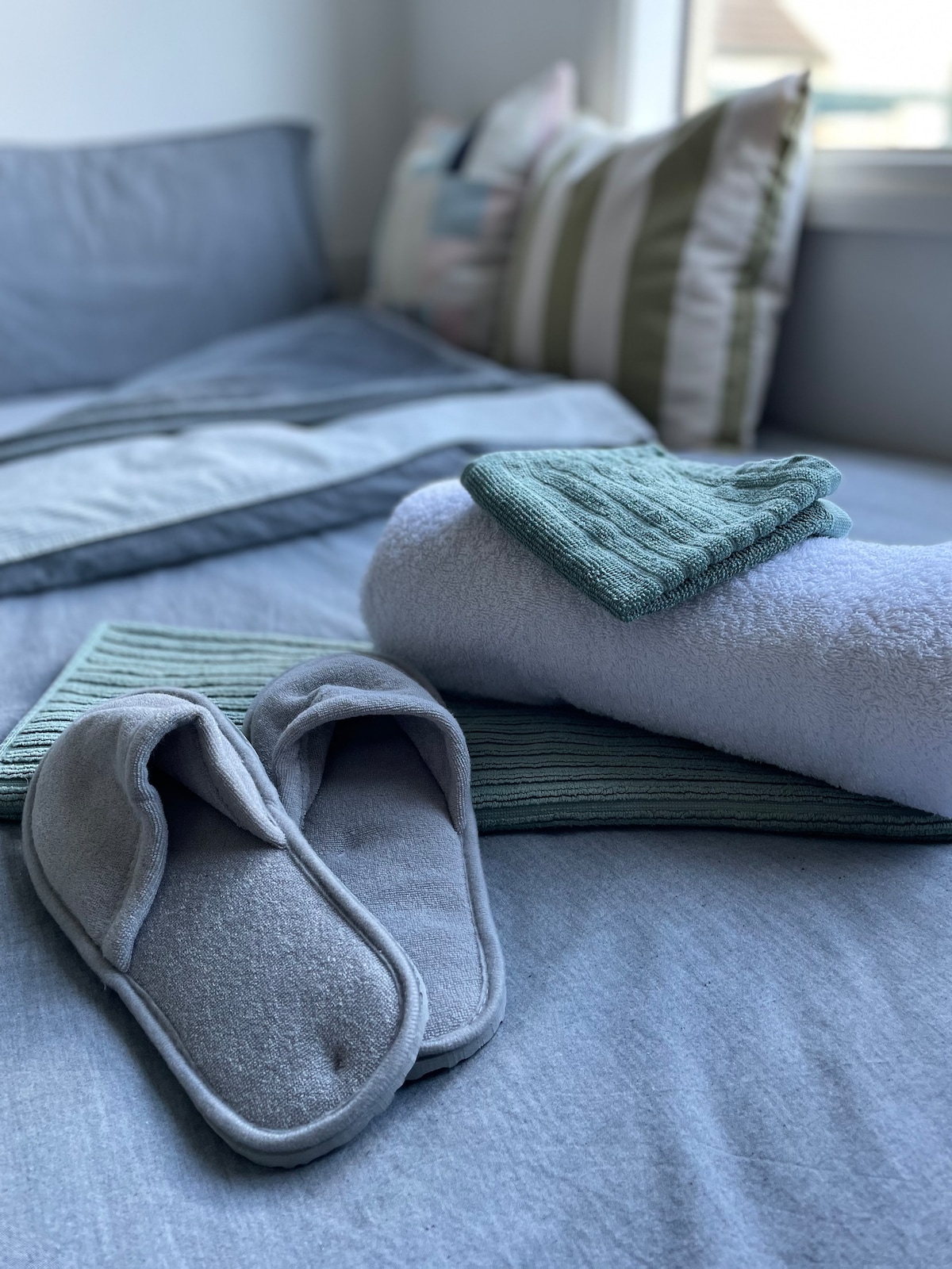 A clean and inviting bed is presented with soft gray linens and accent pillows. Nearby, fresh towels and a pair of plush slippers are neatly arranged, suggesting comfort and convenience for guests.