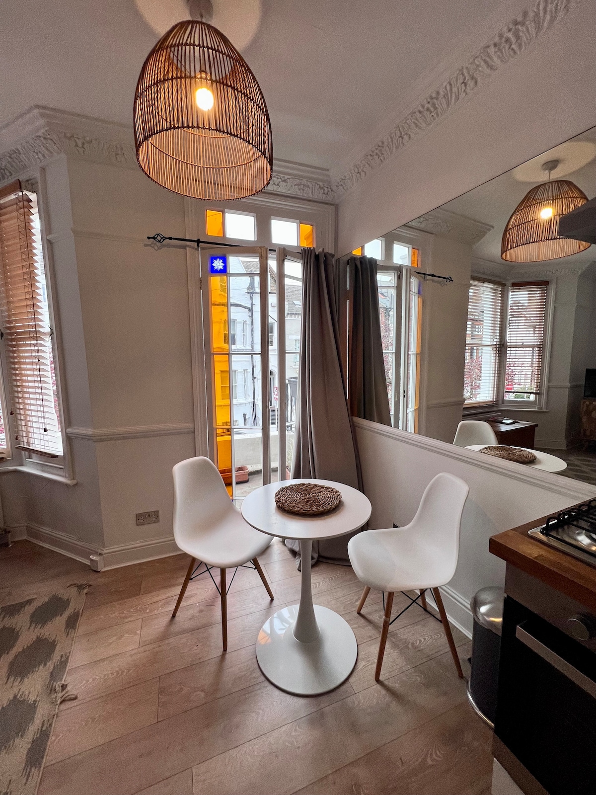 A charming dining area features a round white table surrounded by two modern chairs. Natural light enters through large windows adorned with wooden blinds. The warm glow of woven pendant lights highlights the space, complemented by a partial view of the kitchen with wooden cabinetry.