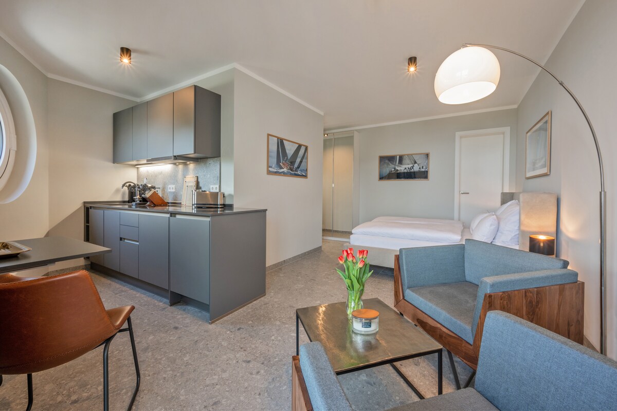An open living and dining area is showcased, featuring a kitchenette with modern appliances. A small table with fresh flowers is positioned near cozy seating, which includes a wooden chair and a soft sofa. A spacious bed is visible in a separate area, ensuring a relaxing ambiance.