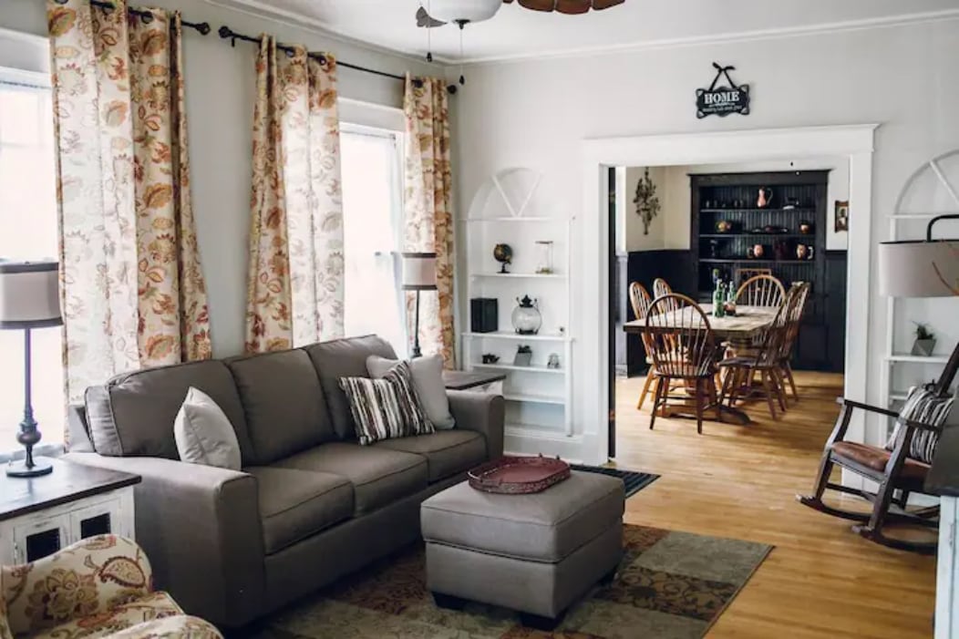 A light-filled living area features a comfortable sofa adorned with decorative pillows, accompanied by an ottoman. Decorative curtains frame the windows. A dining area with wooden chairs is visible through an open space, enhancing the home's inviting layout.