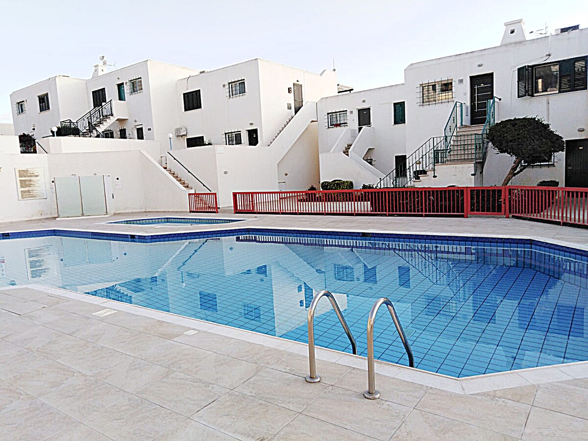 An outdoor swimming pool is depicted, featuring clear blue tiles and metal handrails for easy access. Surrounding the pool, well-maintained white buildings with green accents are visible, as well as a red railing that adds contrast to the scene.