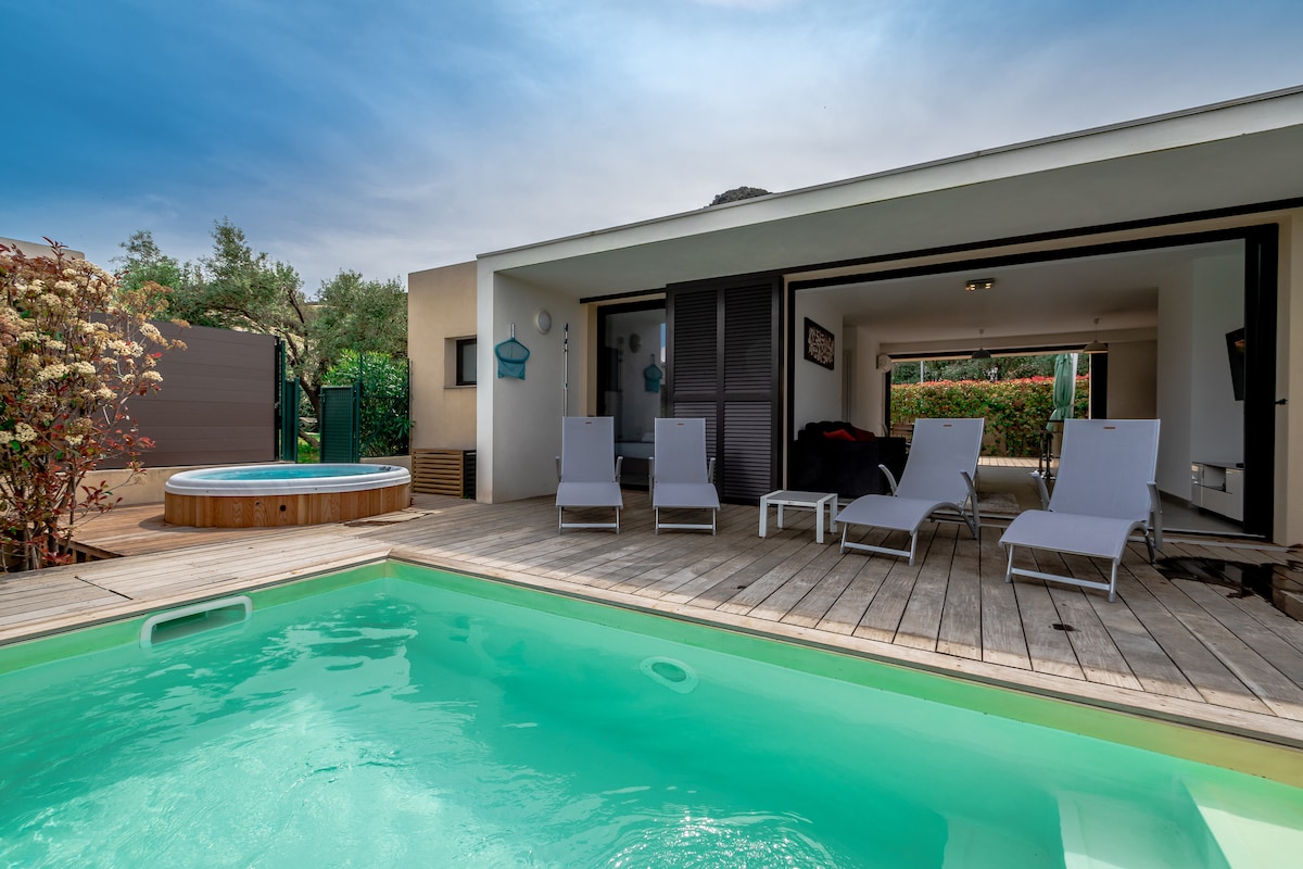 A private wooden deck surrounds a clear turquoise pool, offering ample space for relaxation. Four sun loungers are positioned nearby, while a round hot tub is located beside the pool. Views of lush greenery can be seen in the background.