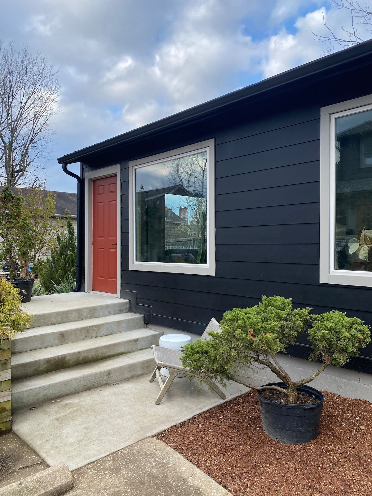 The exterior view showcases a modern cottage with a striking red front door and two large windows. A pathway leads up to the entrance, bordered by landscaped greenery and a potted plant on one side, enhancing the inviting appearance of the space.