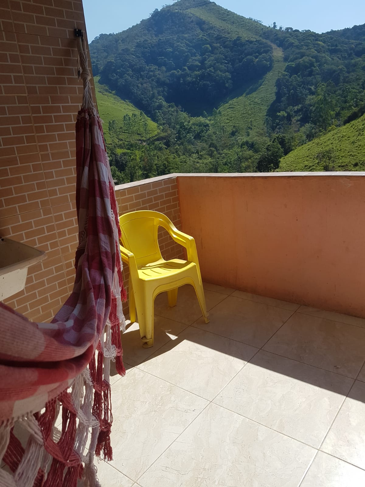 A balcony is shown with a view of rolling green hills. A yellow chair is positioned on the tiled floor, and a soft-textured throw is hung on the railing. The area is bathed in natural light, creating a serene outdoor space.