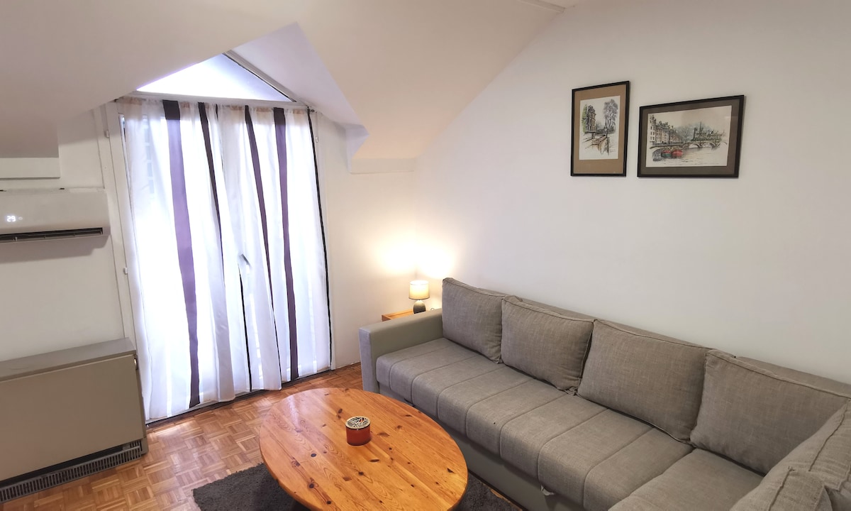 A cozy living area features a light gray sofa arranged around a wooden coffee table. Natural light filters through sheer curtains, illuminating the space. Two framed artworks are displayed on the wall, adding a touch of character to the room.