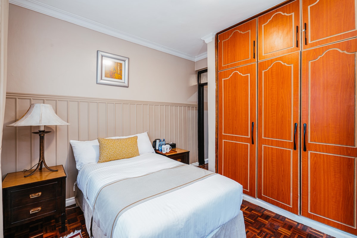 A cozy bedroom features a spacious bed with crisp white linens. A bedside table with a lamp complements the room, while a large wooden wardrobe provides ample storage. Subtle wall paneling adds a touch of elegance to the warm-toned space.