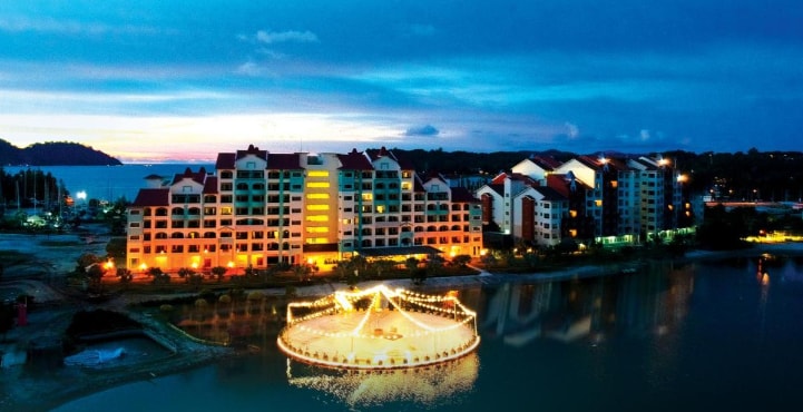 A waterfront view showcases a multi-story building with colorful facades, illuminated softly by exterior lights. A circular platform with lights is positioned in the water, while the horizon features a blend of twilight colors, enhancing the serene atmosphere.