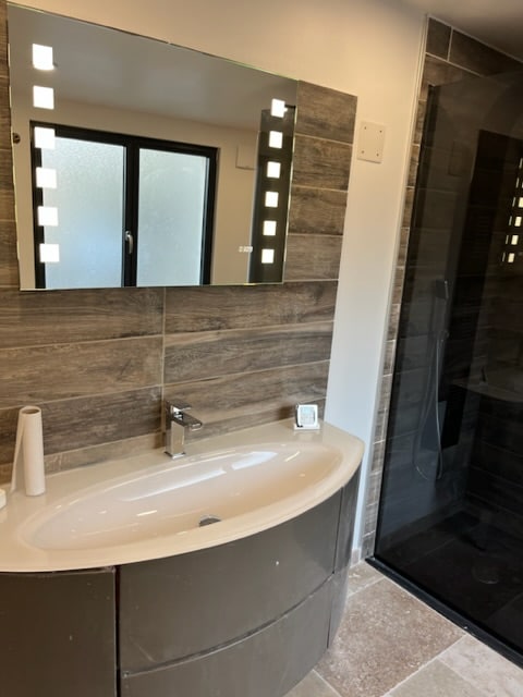 The bathroom features a modern vanity with a smooth, curved sink and a stylish mirror illuminated by led lights. Dark tiled walls complement the sleek shower enclosure, which offers a clear glass door, creating a spacious and inviting atmosphere.