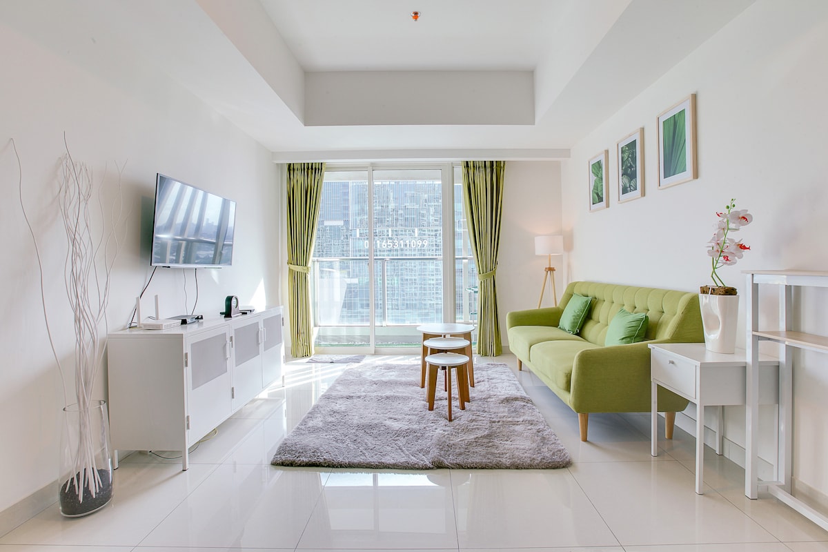 A modern living area is highlighted by a sleek green sofa and a light wood coffee table. Soft rugs cover the glossy floor, reflecting natural light from large glass doors that lead to a balcony. Minimalist décor with framed artwork enhances the space.