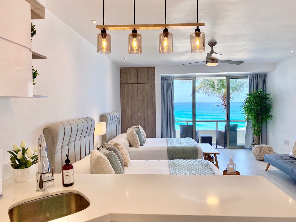 A modern studio features two spacious beds adorned with light-colored linens. A well-equipped kitchen is visible in the foreground, while large glass doors open to a private balcony offering views of the turquoise sea and palm trees swaying in the breeze.